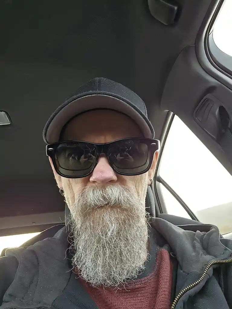 A man with a long white beard, wearing a black baseball cap and large black sunglasses, takes a selfie inside a car. He has…