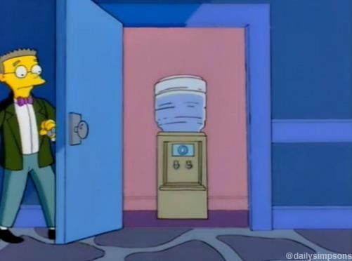 Homer Simpson stands in a doorway looking at a water cooler with a large water bottle on top. The room has blue walls and a…