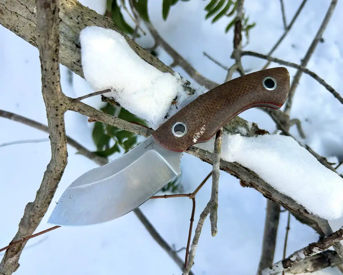 A close-up photo shows a Boker brand knife with a brown textured handle and silver blade resting on snow-covered tree…