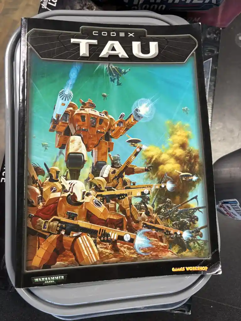 A Warhammer 40,000 Codex: TAU rulebook cover shows orange-armored Tau soldiers and a large mechanized walker firing energy…