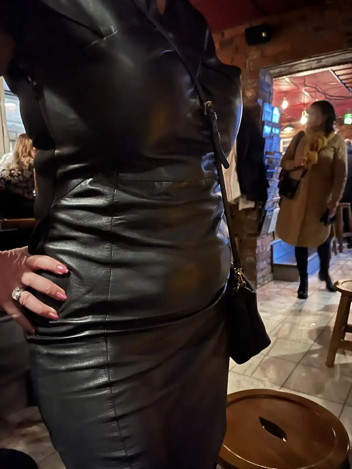 A person wearing a tight black leather dress stands with their hand on their hip, showing the dress's form-fitting…