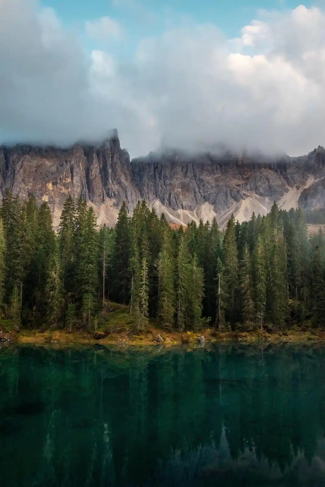 A serene landscape photo shows a calm, emerald-green lake reflecting a dense forest of tall evergreen trees. Behind the…