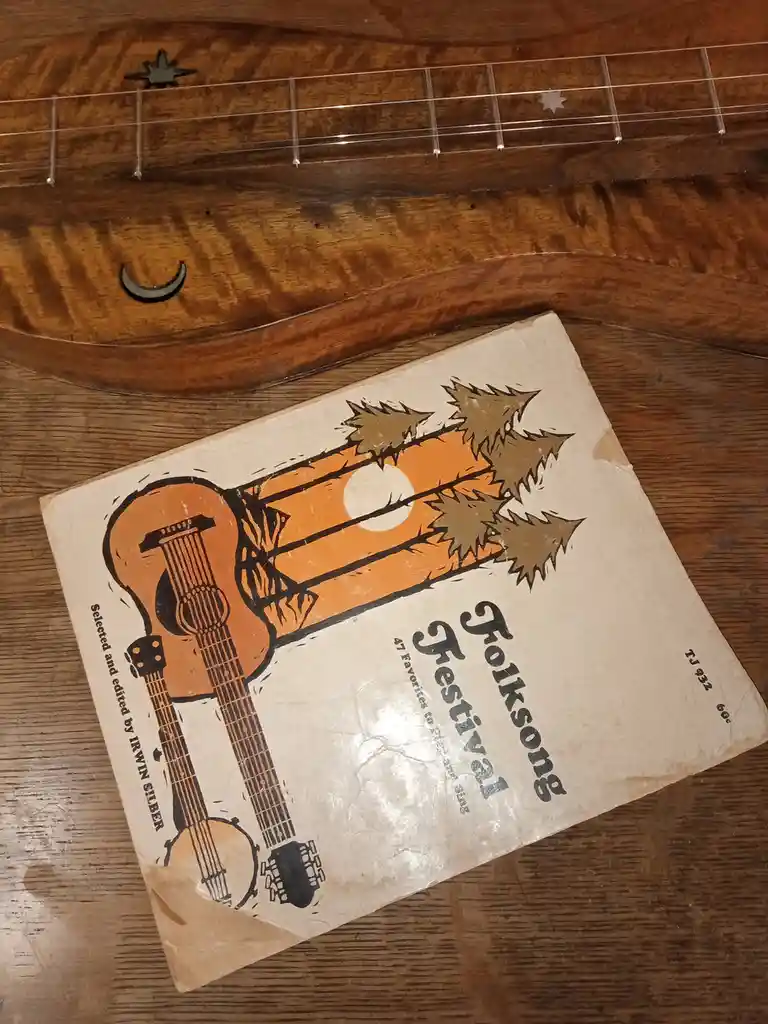 A vintage 'Folksong Festival' songbook, edited by Irwin Silber, lies on a wooden surface next to the neck of a wooden…