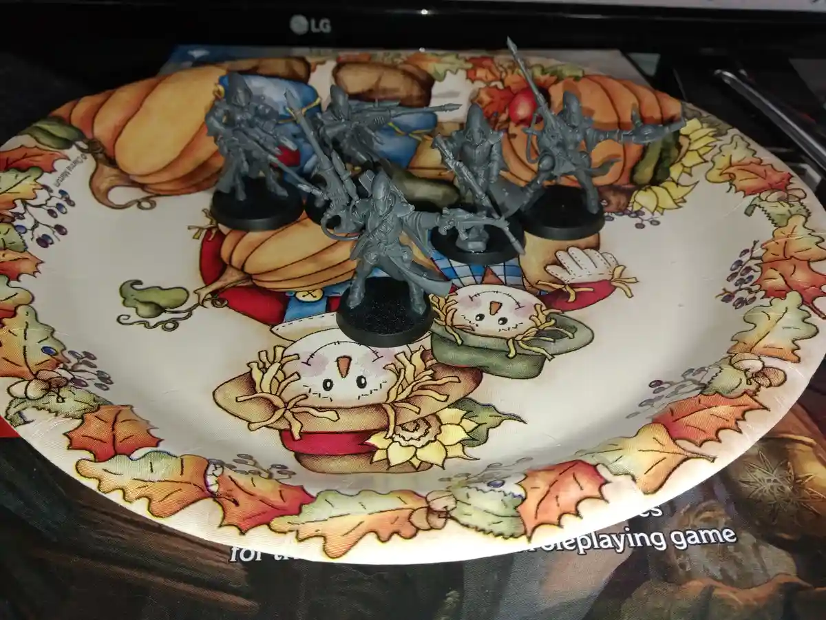 Several gray, unpainted fantasy miniatures of armed warriors are arranged on a festive Thanksgiving-themed plate featuring…