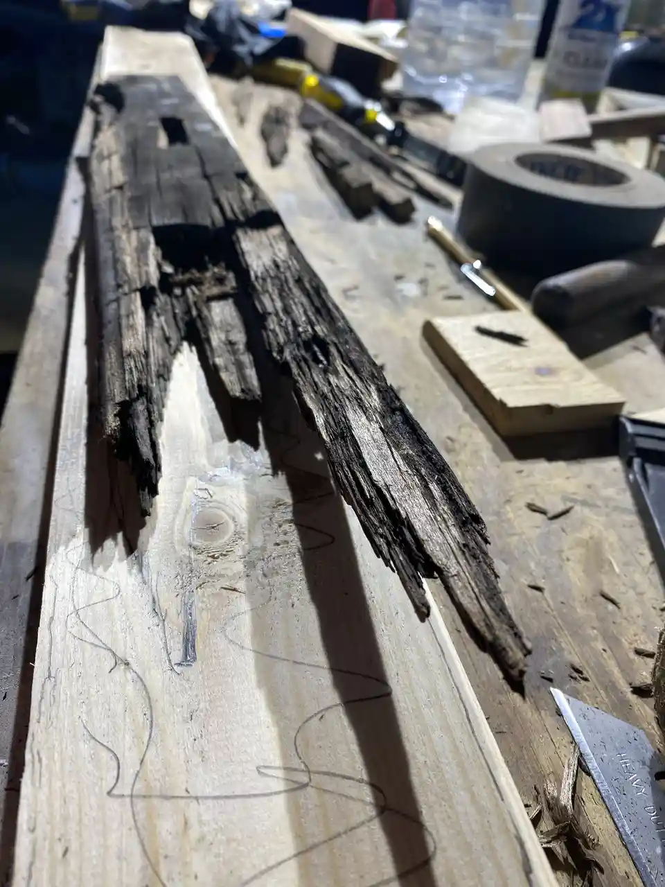 A close-up photo shows a piece of dark, charred, and splintered wood resting on a lighter, freshly marked wooden plank on a…