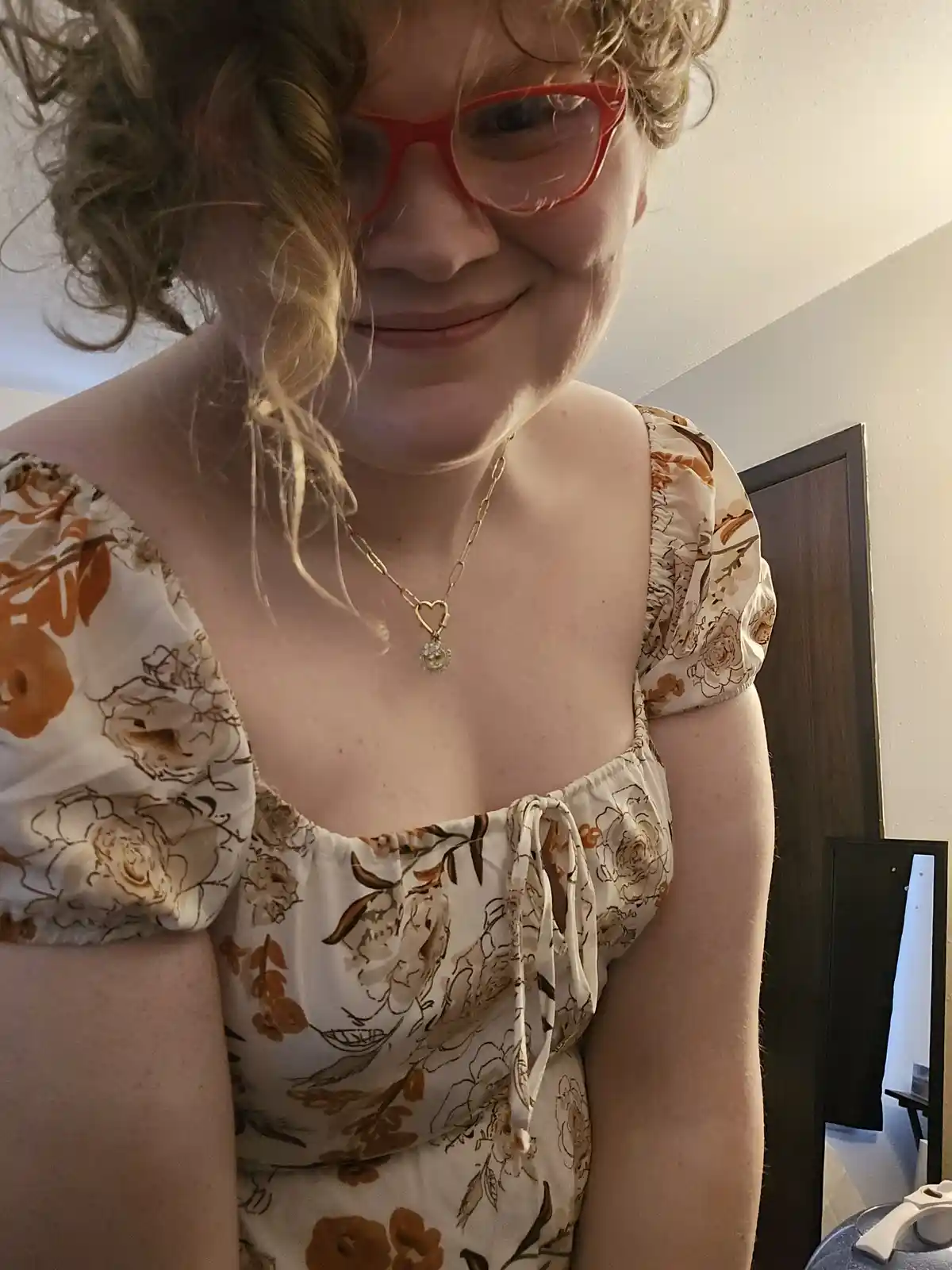 A person with curly blonde hair and red glasses smiles while wearing a floral top and a gold heart-shaped necklace. The…