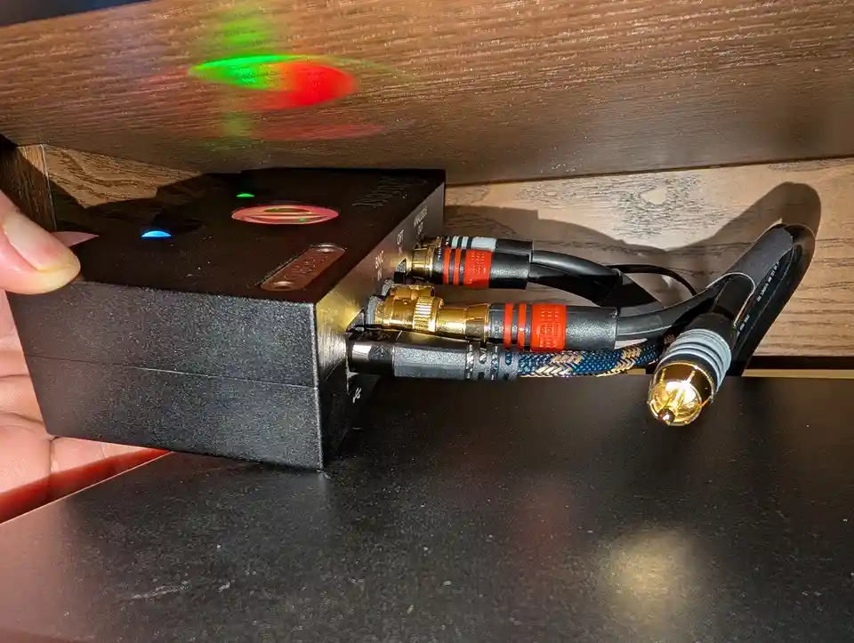 A hand holds a small black audio device with glowing indicator lights, connected to thick, expensive-looking cables with…