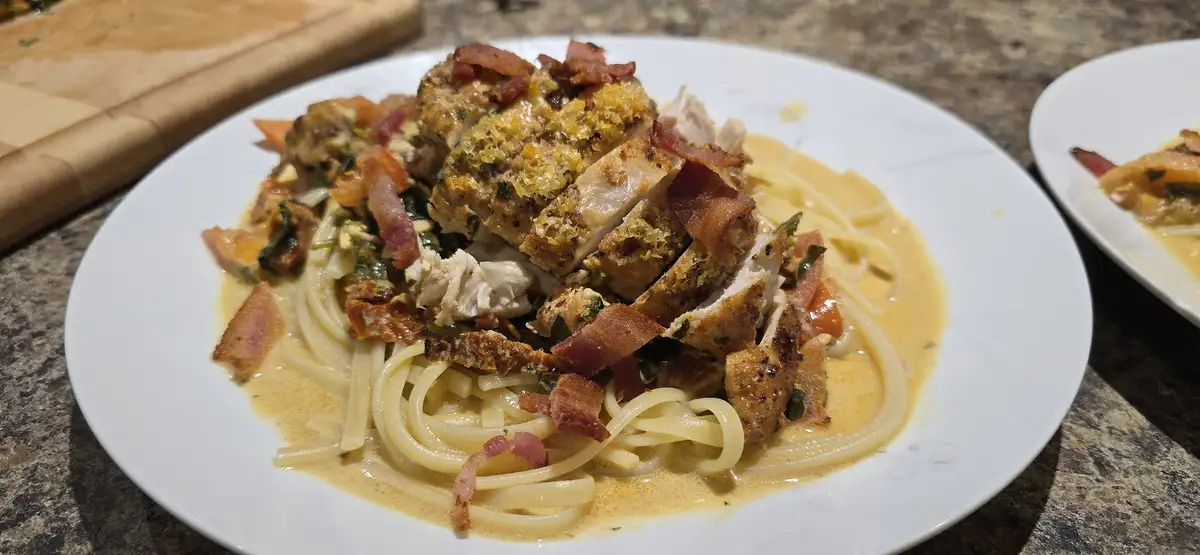 A close-up photo of a white plate holding pasta with creamy sauce, sliced breaded chicken, and crispy bacon pieces. The dish…