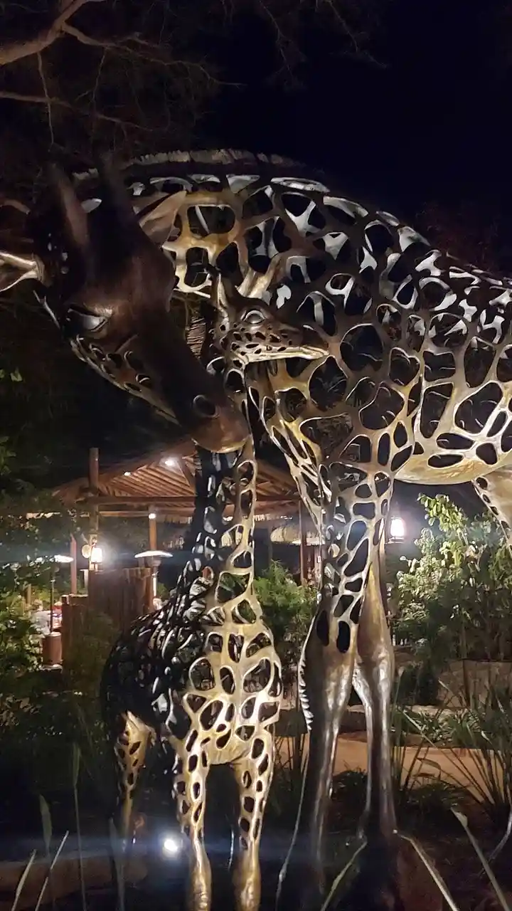 Two large, metallic giraffe sculptures stand close together at night, their bodies featuring a distinctive perforated…