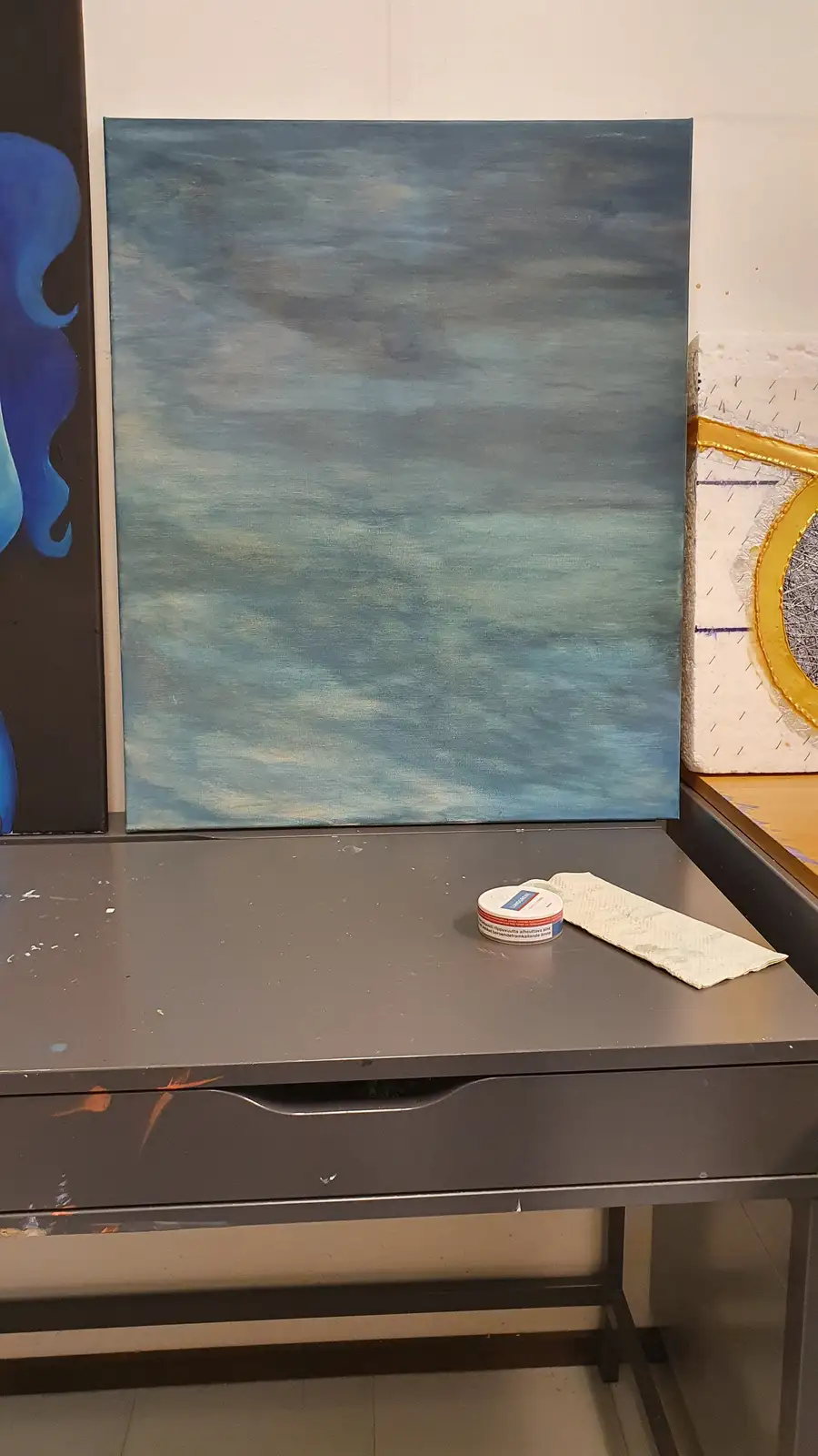 A large, unfinished painting of a cloudy sky leans against a wall in an art studio, with a small tin of Vaseline and a paper…
