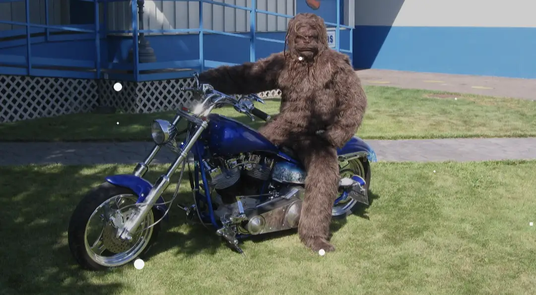 A person in a full-body brown Bigfoot costume is seated on a blue custom motorcycle parked on grass. The motorcycle has…