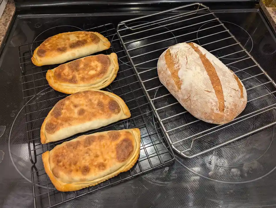 Four golden-brown empanadas sit on a cooling rack next to a rustic, flour-dusted loaf of bread, both resting on a black…