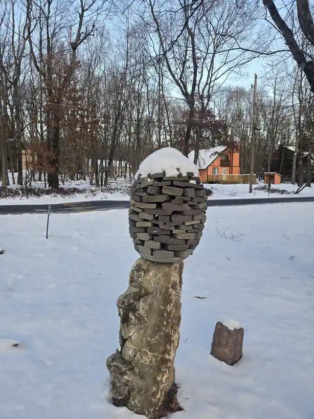 A snow-covered stone sculpture shaped like a human head with a stone body stands in a snowy yard, with bare trees and a road…