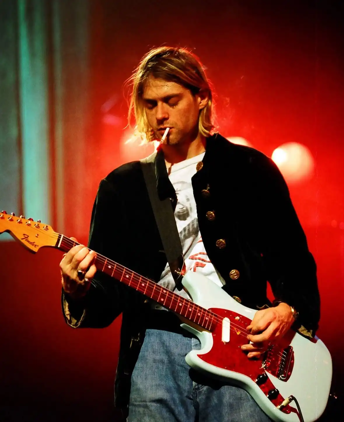 A man with long blonde hair, wearing a black jacket over a graphic t-shirt and jeans, plays a light blue and red Fender…