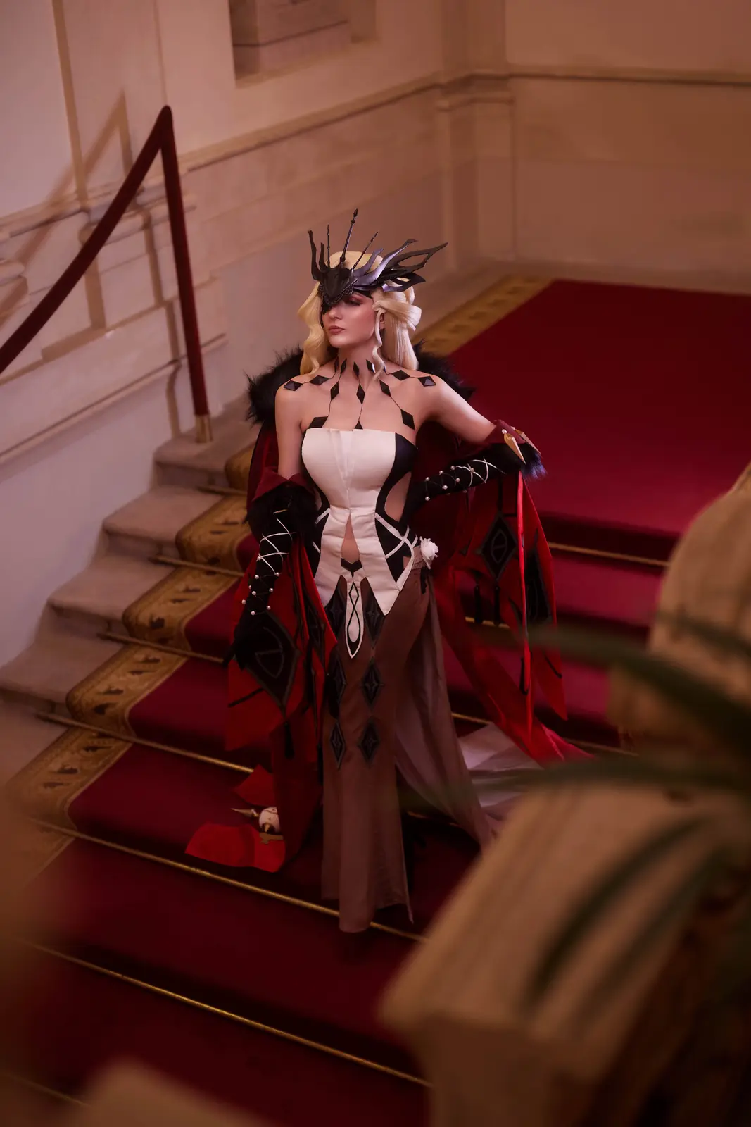 A cosplayer in an elaborate black, white, and red costume with a dramatic crown and cape stands poised on a grand staircase…