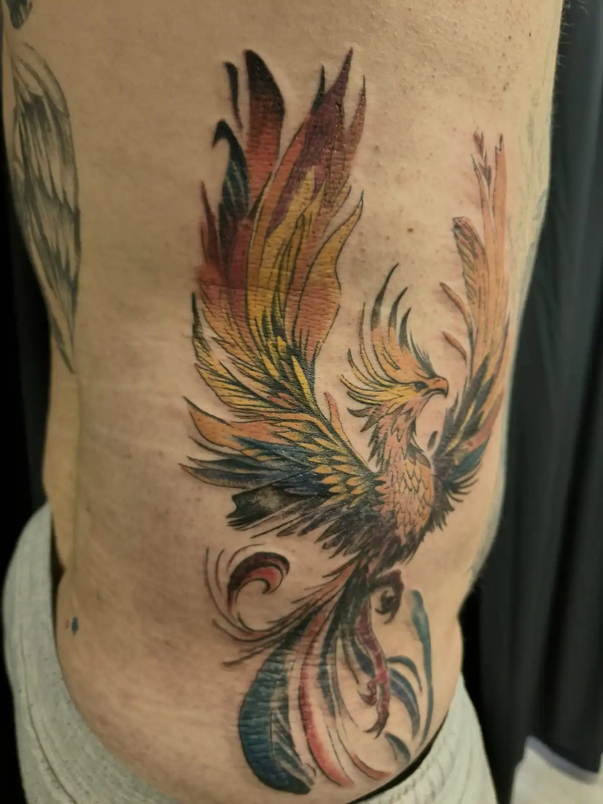 A close-up photo shows a colorful phoenix tattoo on a person's side, featuring vibrant orange, yellow, and blue feathers…