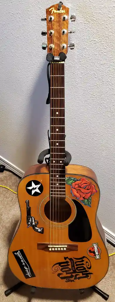 A Fender acoustic guitar stands upright on a stand, heavily decorated with stickers and decals including a rose, a revolver…
