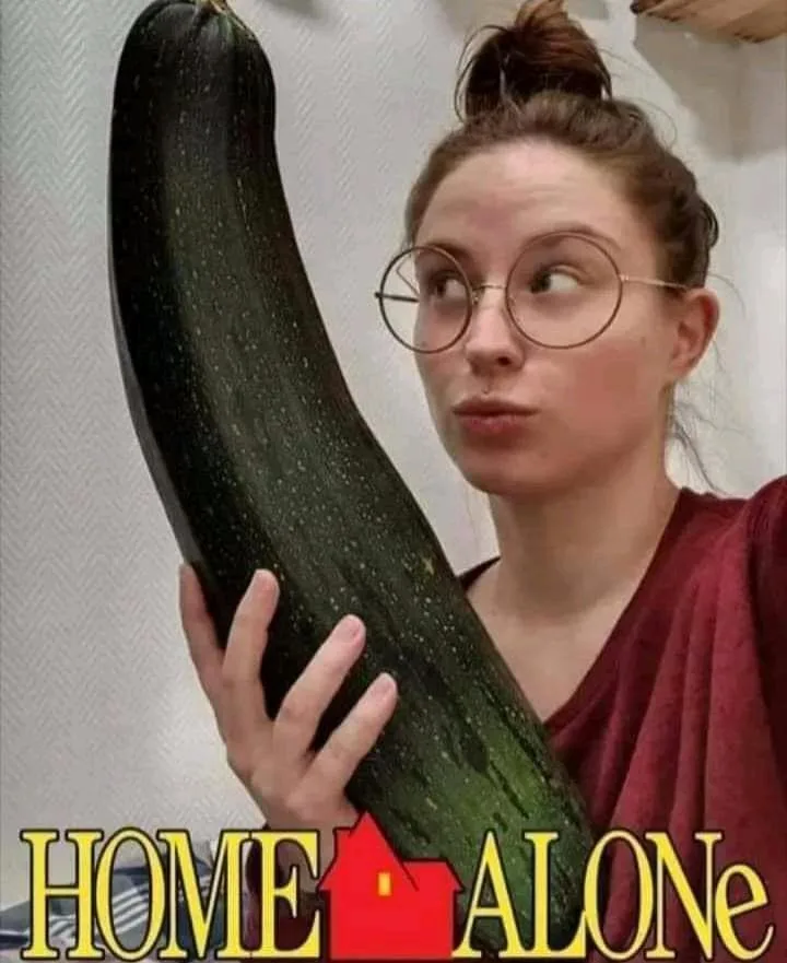 A woman with glasses and her hair in a bun holds an extremely large, dark green zucchini. She looks sideways with a…
