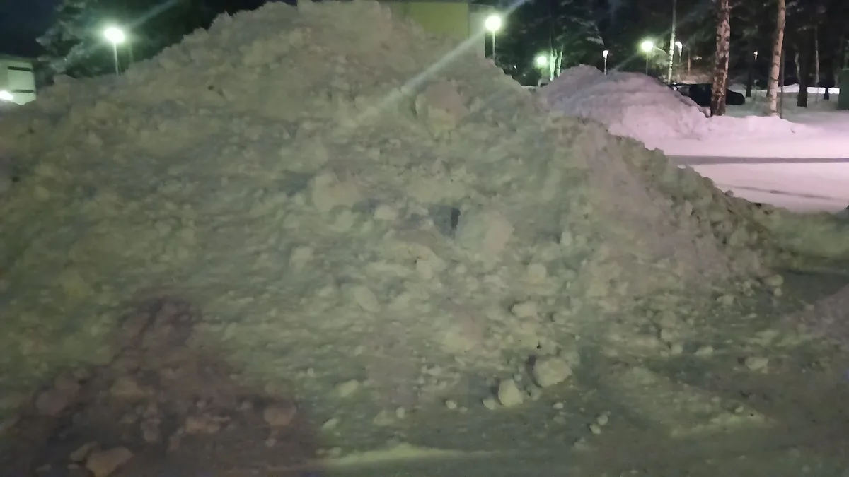 A large, uneven pile of dirty snow dominates a nighttime street scene, illuminated by streetlights, with trees and a…