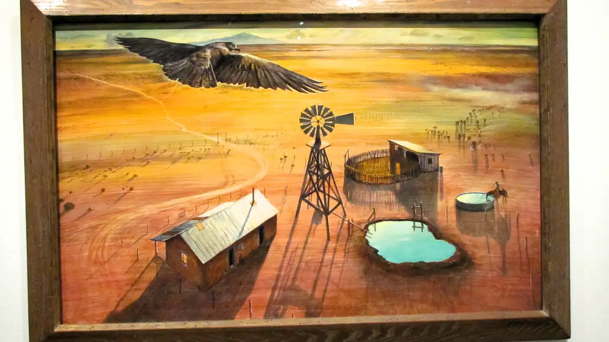A framed painting depicts a rural landscape with a farmhouse, windmill, fenced paddock, and a large bird of prey flying…