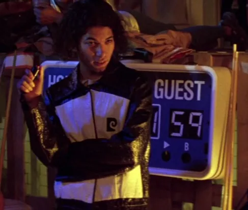 A person with curly hair wearing a black and white jacket stands beside a digital scoreboard labeled 'GUEST' showing the…
