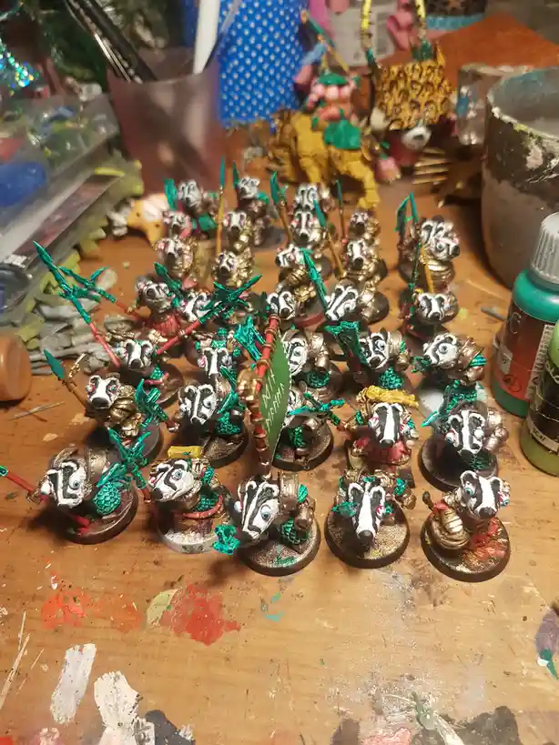 A cluttered, paint-splattered workbench holds dozens of intricately painted miniature badger warriors, each holding a spear…