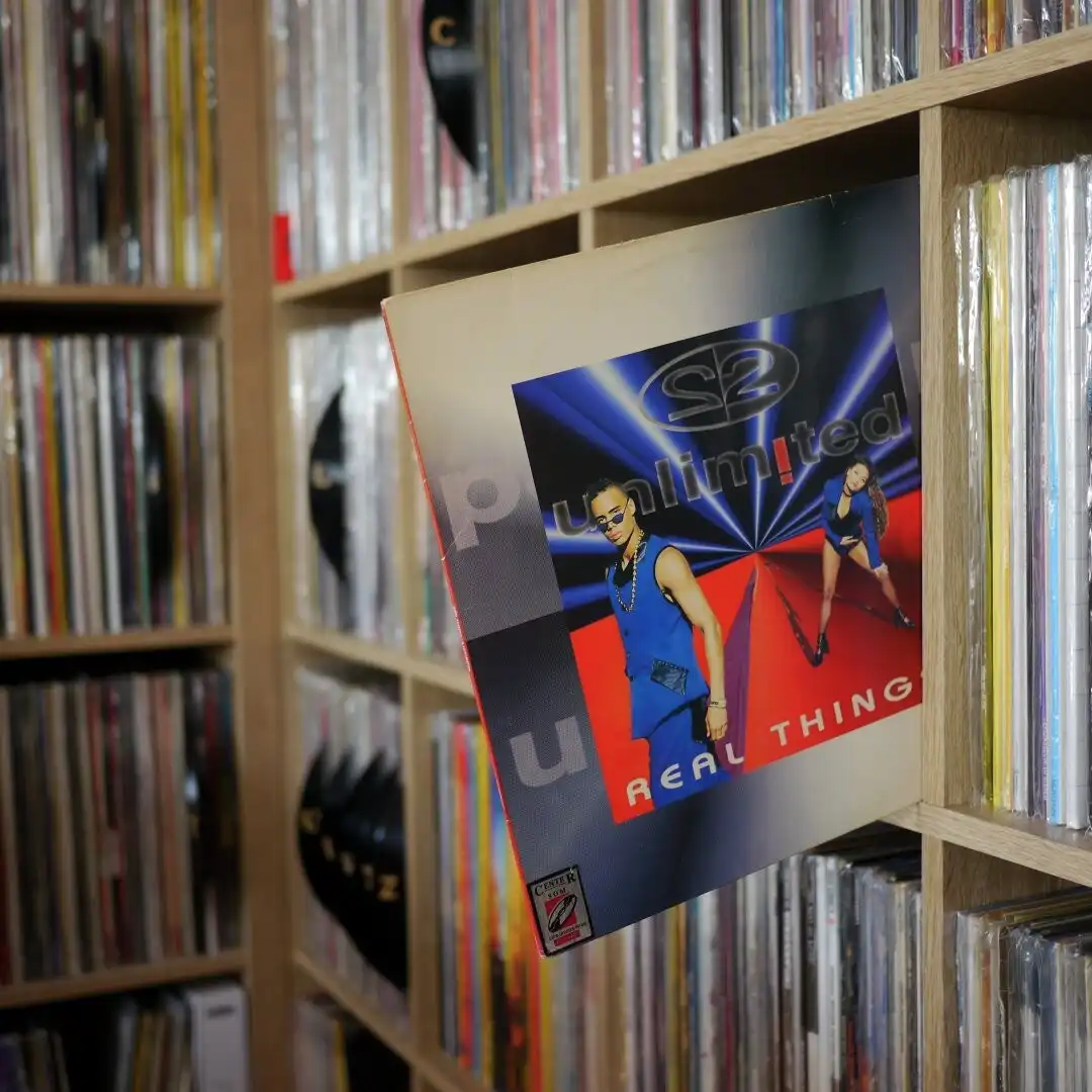 A close-up photo of a CD case for the 1990s group S2, titled 'Unlimited Real Thing', leaning out from a wooden shelf filled…
