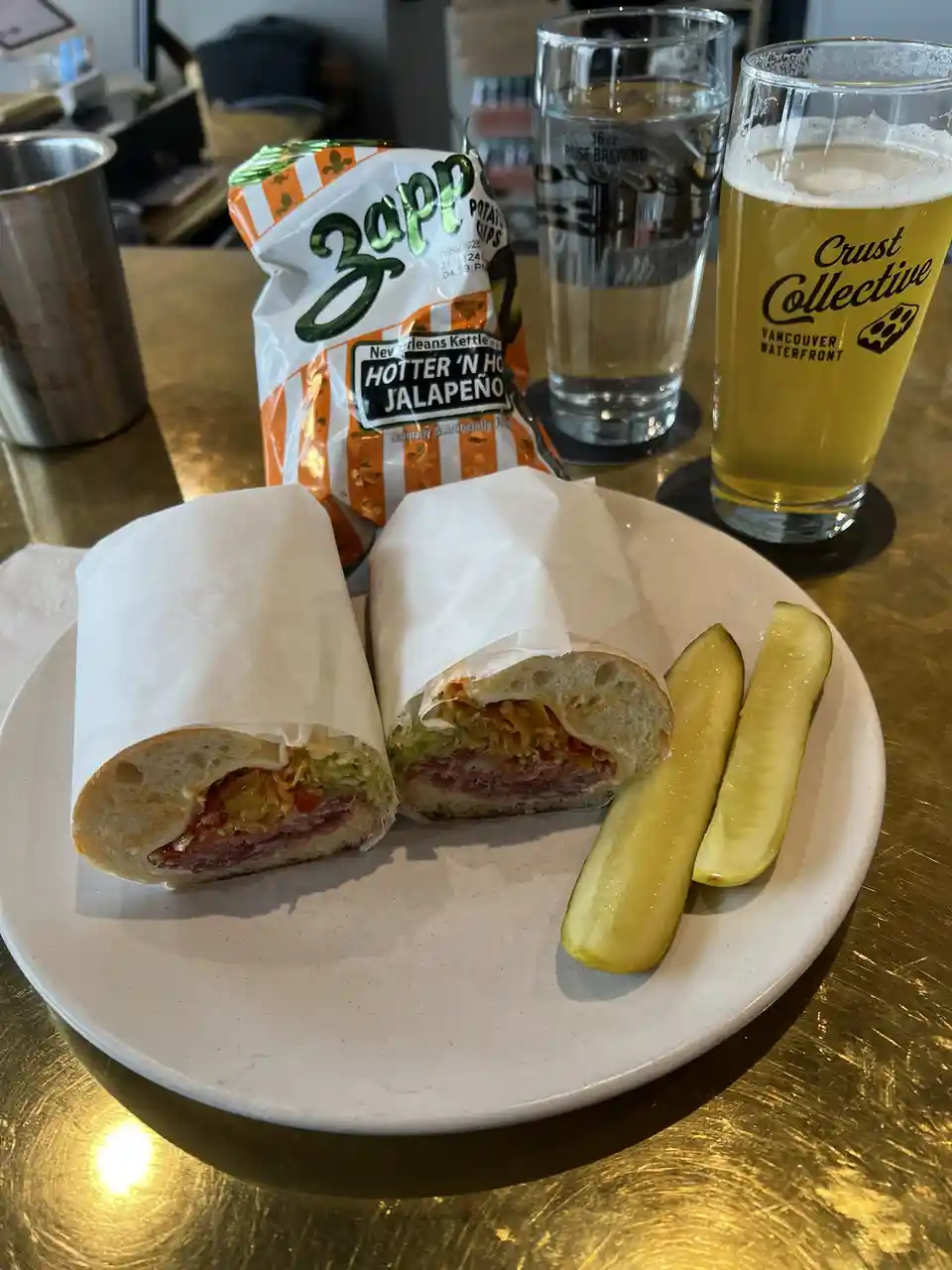 A plate with a sandwich cut in half, revealing meat, cheese, and veggies, accompanied by two pickle slices. Behind it is a…