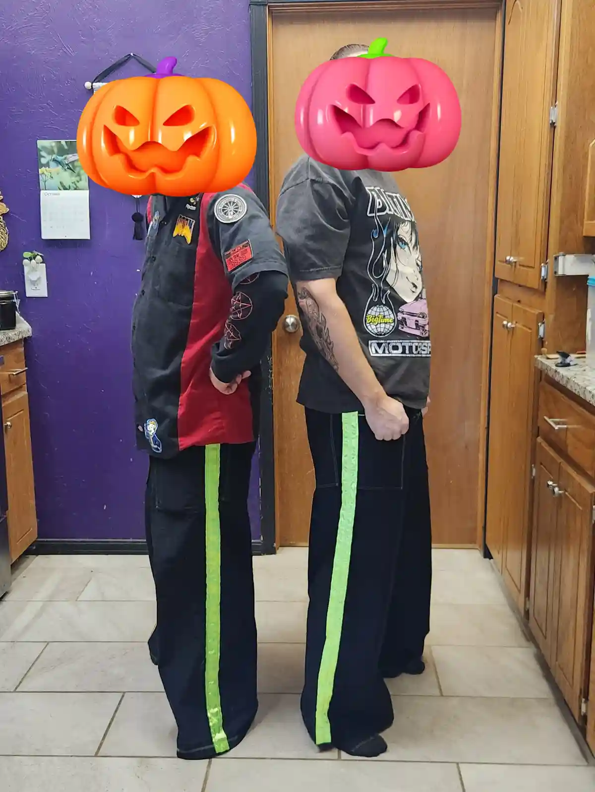Two people stand back-to-back in a kitchen, both wearing black pants with bright green stripes down the sides. Their faces…