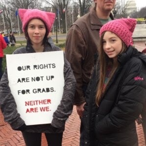 Two young girls wearing pink cat-ear hats stand outdoors holding a white protest sign that reads 'OUR RIGHTS ARE NOT UP FOR…