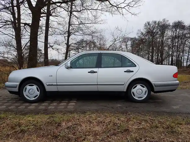 A silver Mercedes-Benz sedan is parked on a cobblestone surface with bare trees in the background under an overcast sky. The…