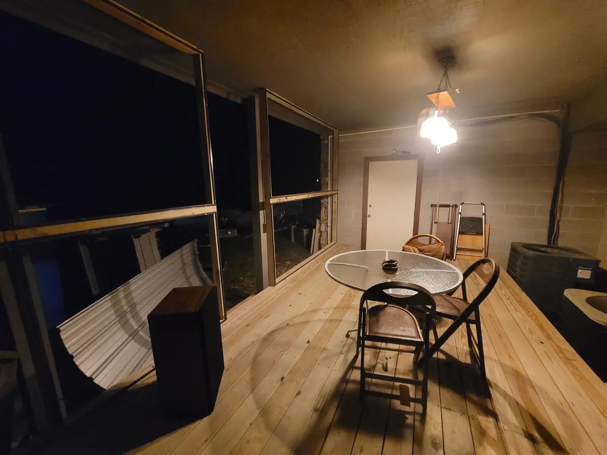 A dimly lit, enclosed porch at night with wooden flooring, a glass-top table with two chairs, and large windows showing…