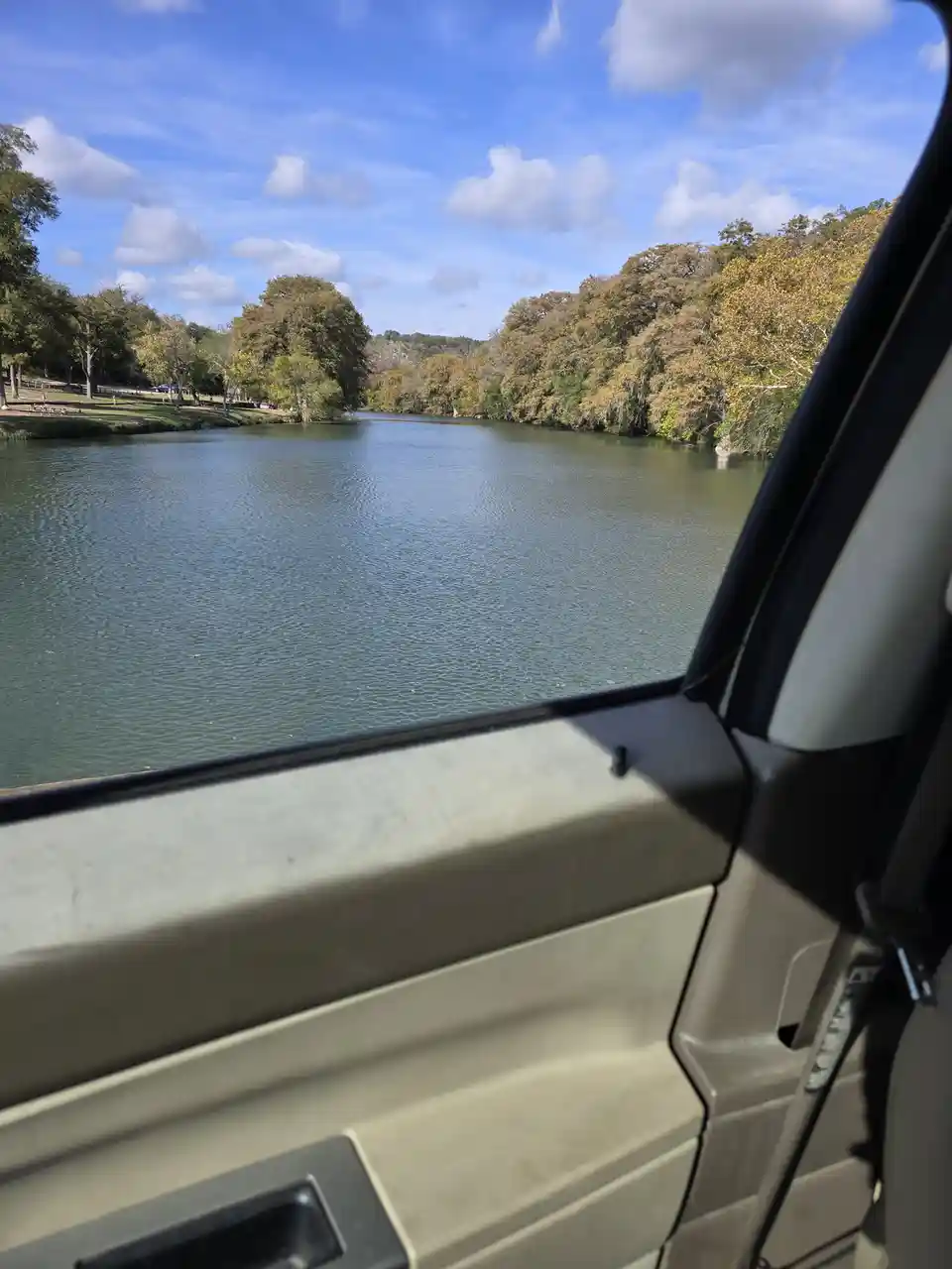 A photo taken from inside a vehicle looking out the window at a calm river surrounded by trees under a partly cloudy sky…