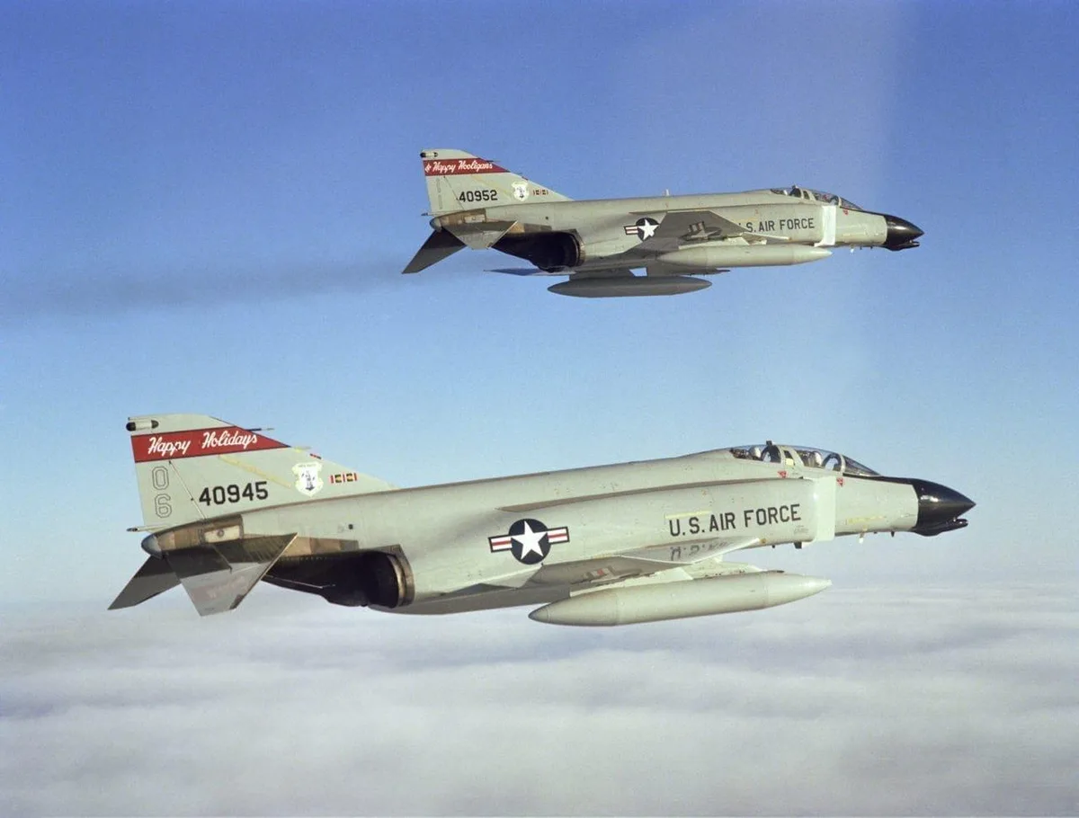 Two U.S. Air Force F-4 Phantom II fighter jets fly in formation above clouds, both with 'Happy Holidays' written in red on…