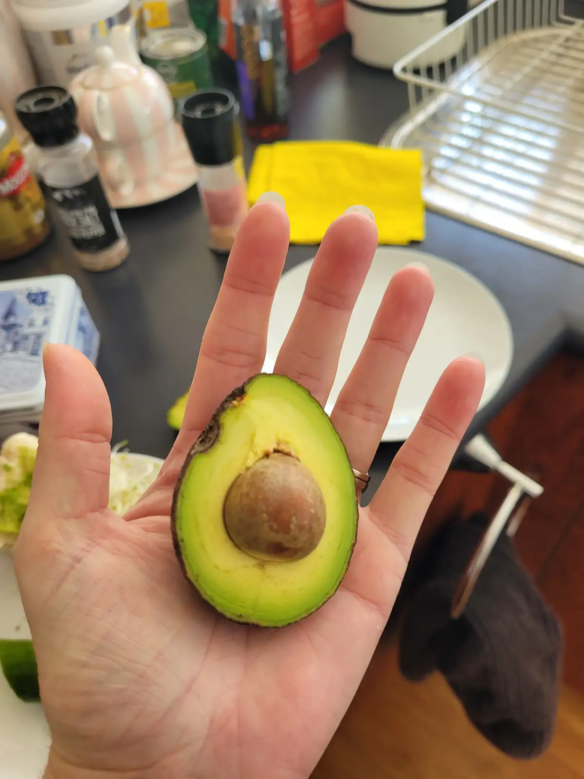 A person holds a halved avocado in their open palm, displaying its large brown pit. The background shows a kitchen counter…