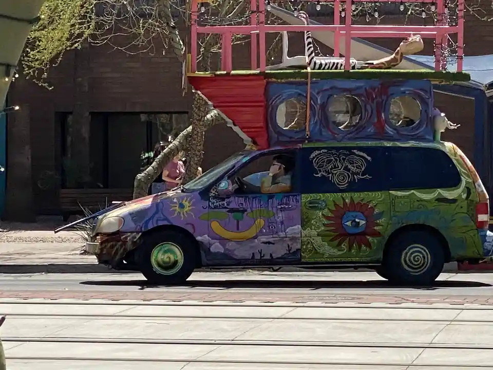 A heavily customized, psychedelic minivan with vibrant, colorful artwork including suns, spirals, and a sun with an eye, is…