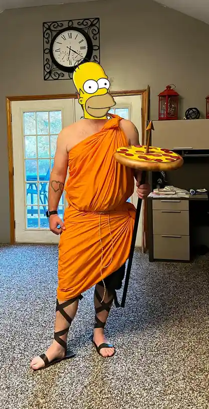 A person stands indoors wearing an orange toga, black strappy sandals, and holding a large pizza on a pole like a spear…