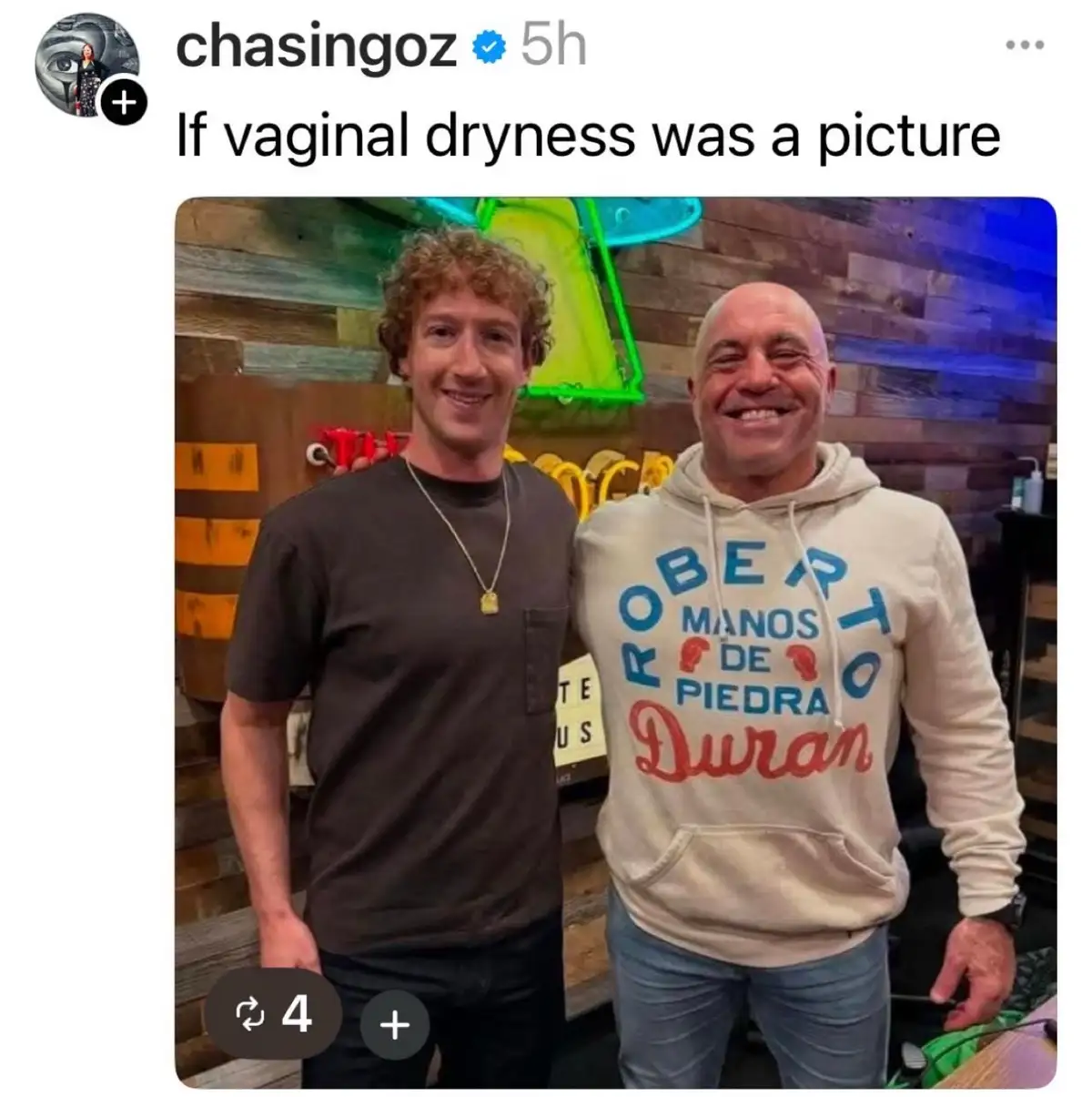 Two men are smiling and posing for a photo. The man on the left has curly hair and wears a dark t-shirt with a gold pendant…