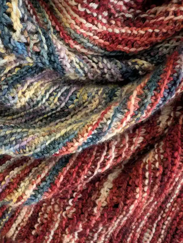 A close-up photo of a hand-knitted scarf with a variegated, multicolored yarn featuring shades of red, orange, yellow, blue…