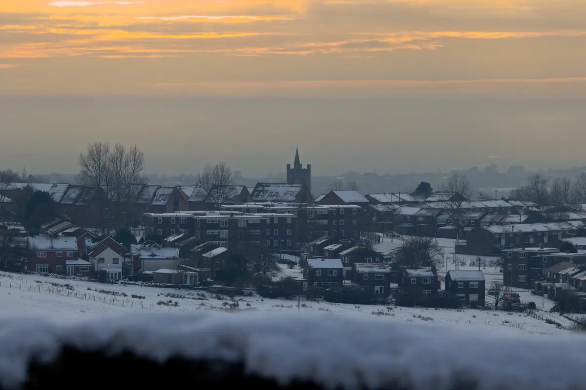 A scenic winter view of a residential neighborhood with snow-covered rooftops and fields under a hazy, orange-tinged sky. A…