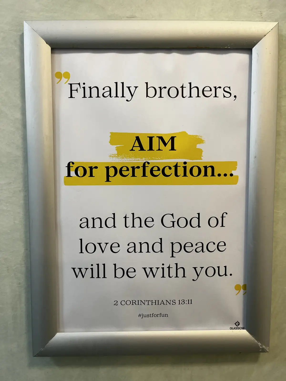 A framed religious quote from 2 Corinthians 13:11 is displayed on a wall, with the word 'AIM' humorously highlighted in…
