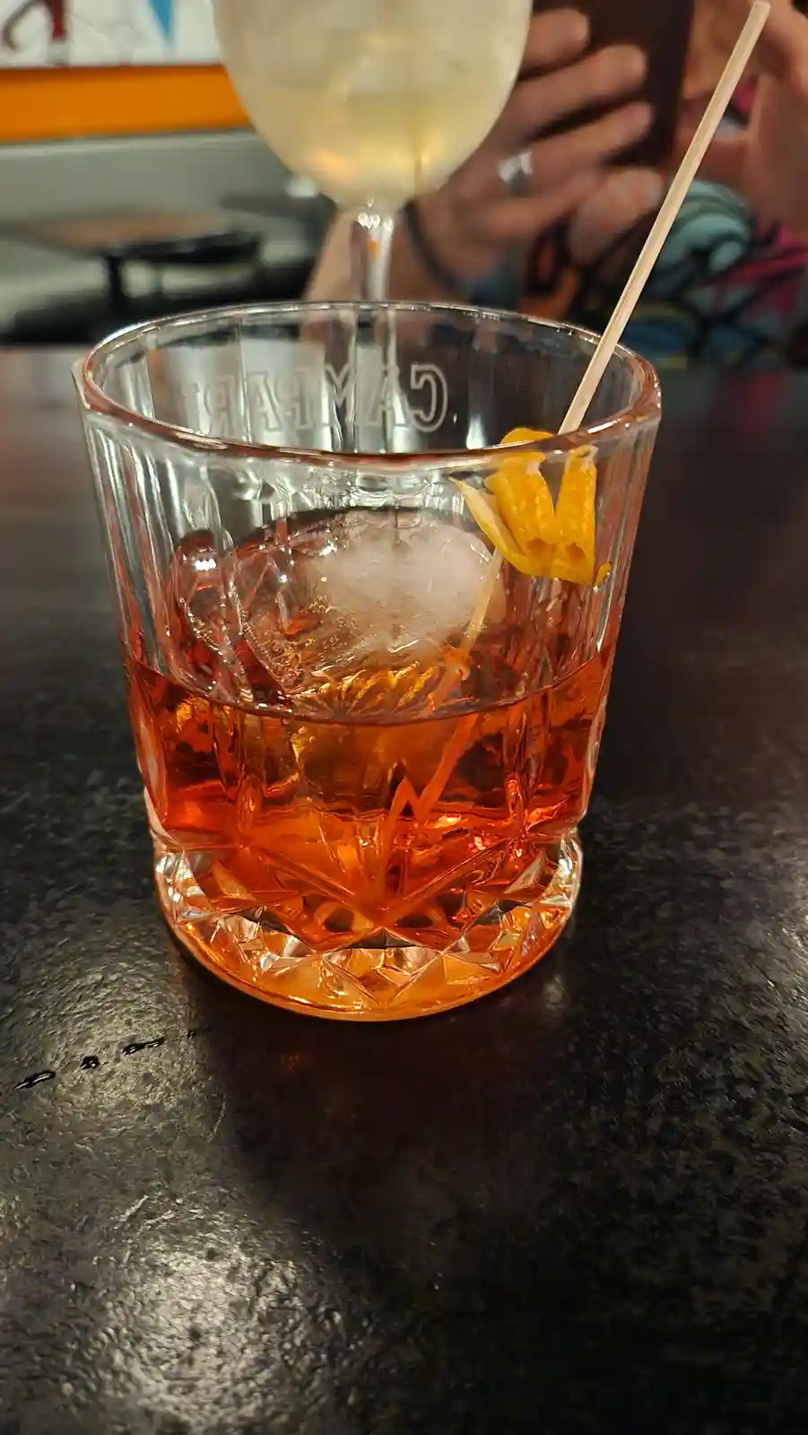 A close-up photo of a Campari cocktail in a faceted glass with ice and an orange peel garnish, sitting on a dark bar…