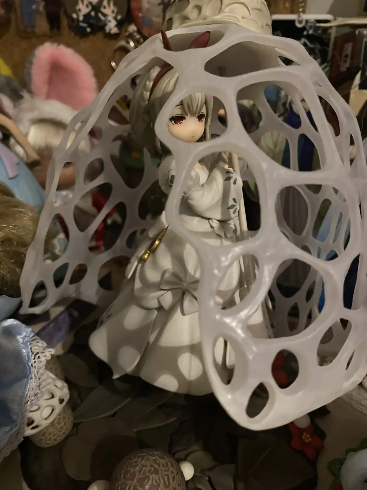A close-up photo shows a white anime-style figurine with red eyes and a white dress, partially obscured by a large, white…
