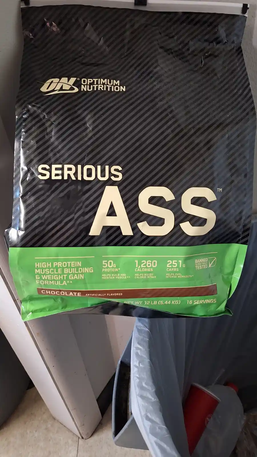 A large black bag of Optimum Nutrition protein powder, labeled 'SERIOUS ASS', is hanging above a trash can. The bag's…