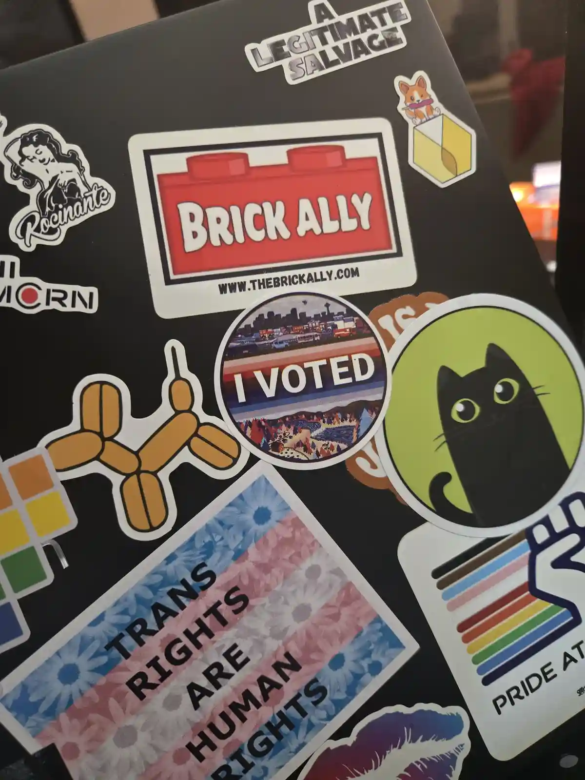 A close-up of a black surface covered with various colorful stickers, including 'BRICK ALLY', 'I VOTED', 'TRANS RIGHTS ARE…