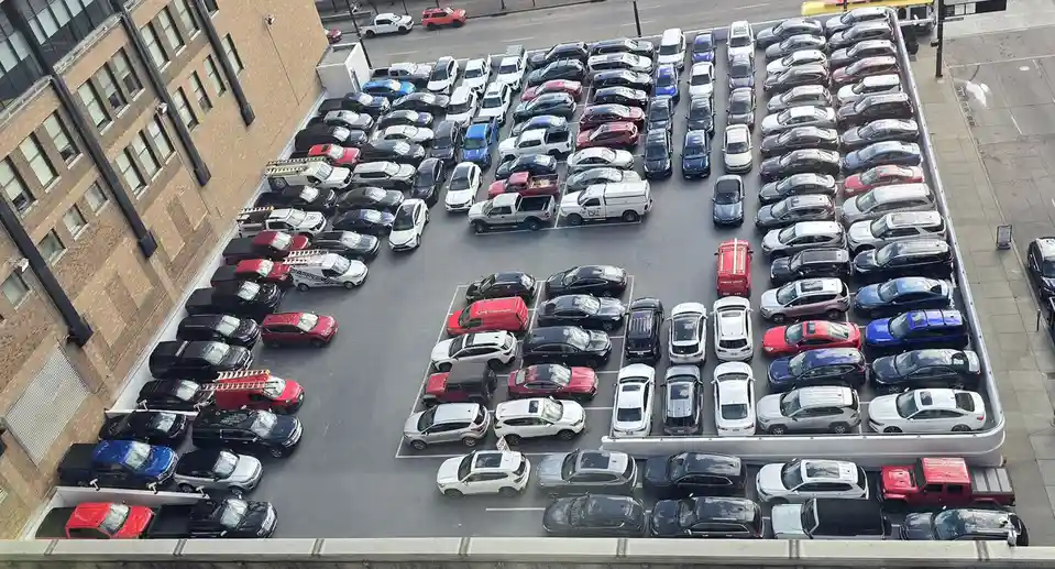 An aerial view of a densely packed rooftop parking lot filled with numerous cars of various colors and models, adjacent to a…