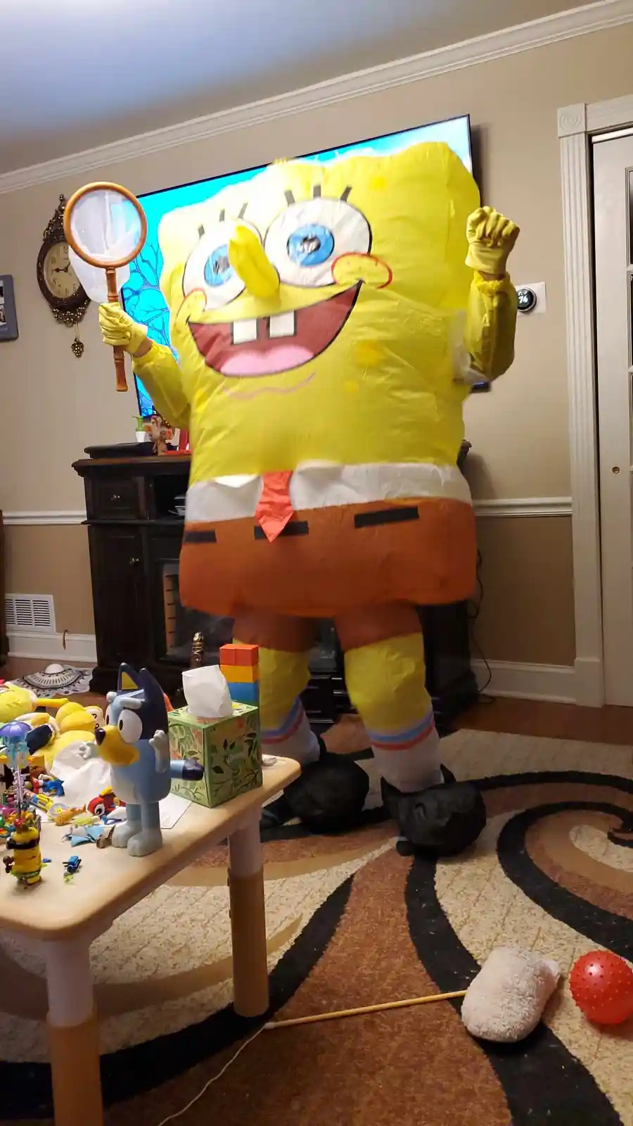 A person is wearing a large, inflatable SpongeBob SquarePants costume, standing in a living room. The character is holding a…
