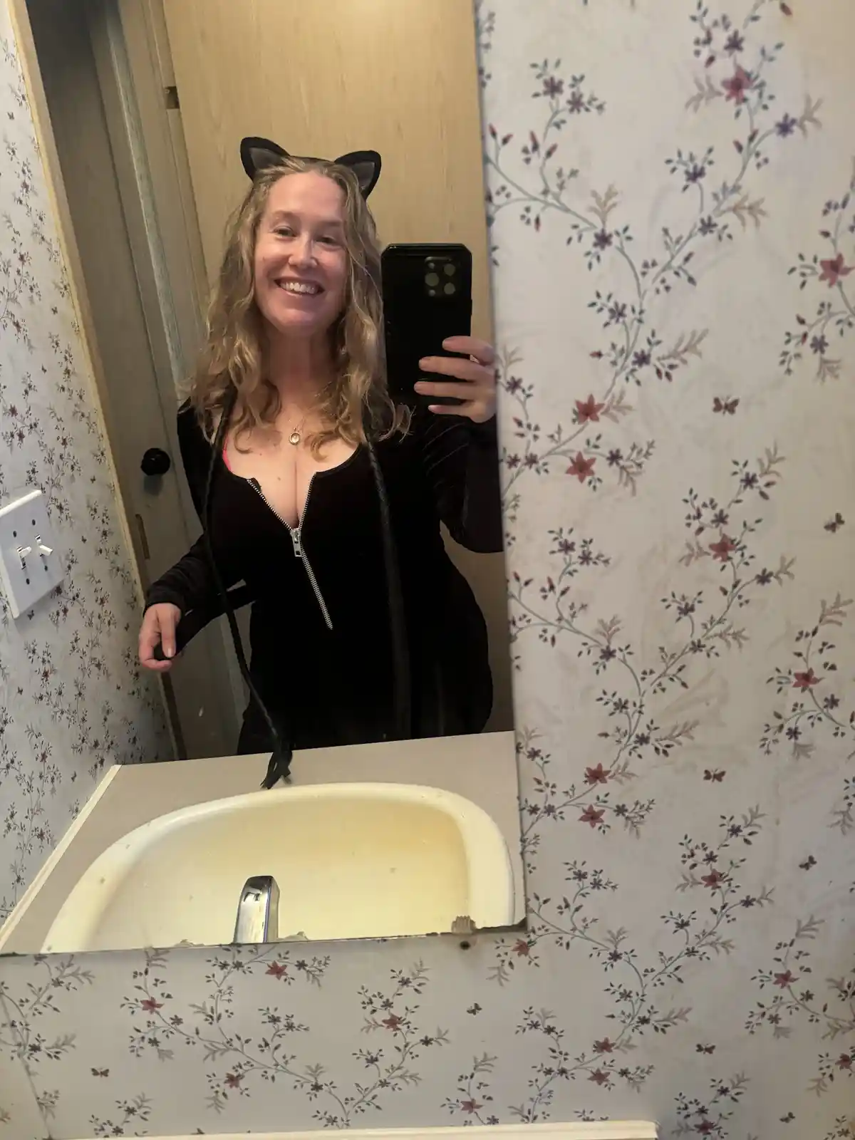 A woman with blonde hair wearing black cat ears and a black zip-up costume with a tail, takes a mirror selfie in a bathroom…