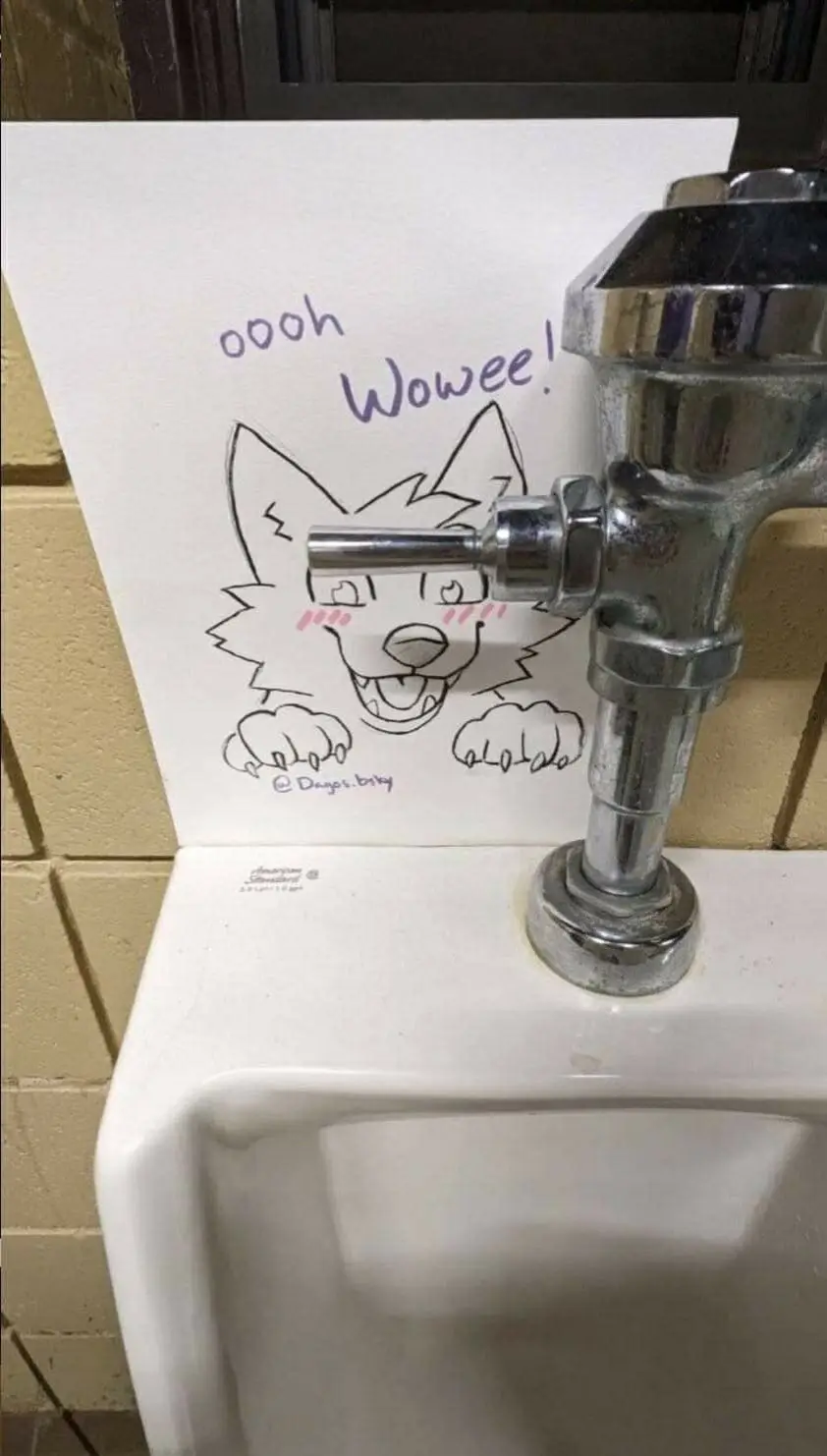A hand-drawn cartoon wolf with blush marks and an excited expression is taped to the wall behind a urinal, with its drawn…