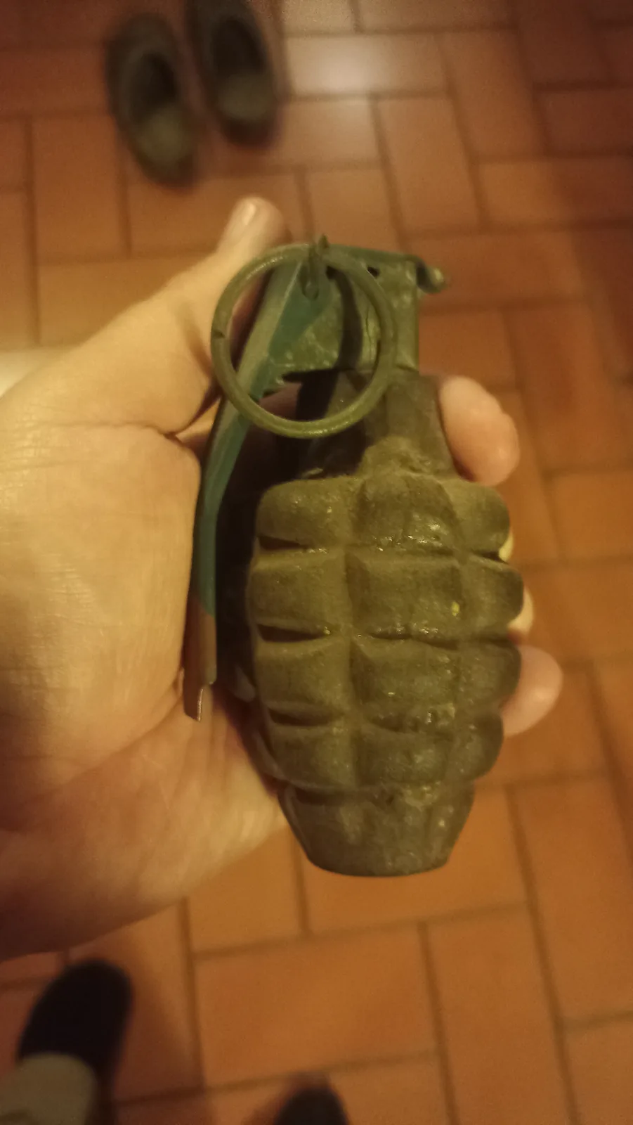 A hand holds a green, textured hand grenade with a ring and safety lever, against a blurred background of tiled floor and…